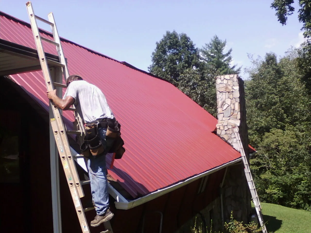 Roofer on a ladder beside a red metal roof and stone chimney — Soffit & Fascia Repair by Roofing Brothers in Trade